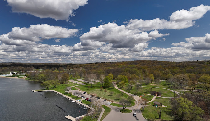 Aerial view of a park in Michigan with winding pathways and a pier extending into a lake, surrounded by lush greenery and puffy clouds above.
