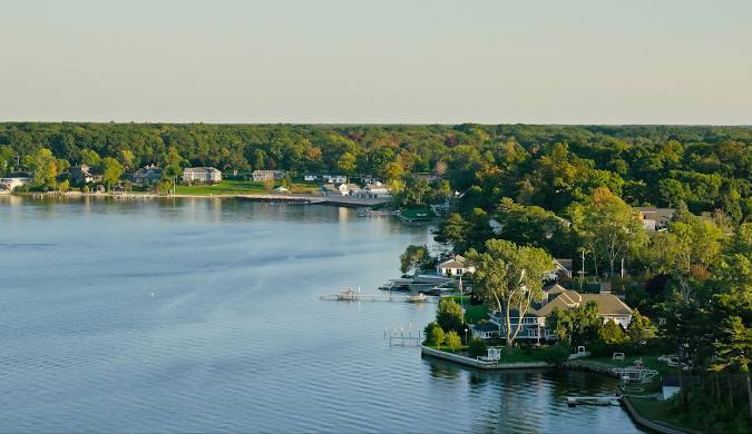 Aerial view of a calm lake in Michigan with residential houses and docks along the shoreline, surrounded by lush green trees under a clear blue sky.