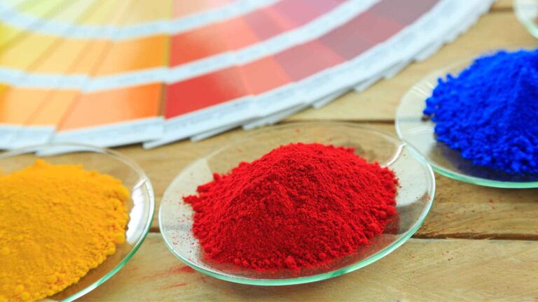 Bowls of yellow, red, and blue powder pigments are arranged on a wooden surface, with a color palette in the background.