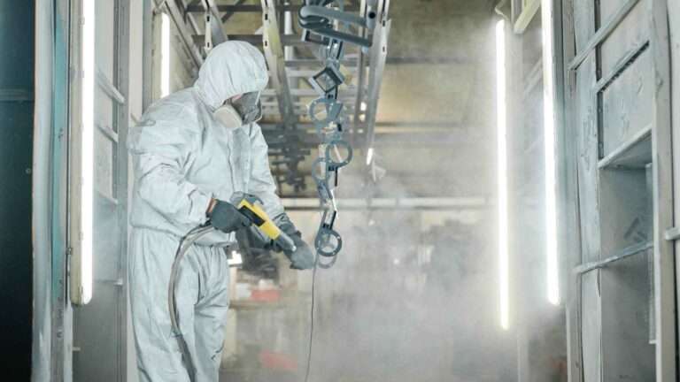 A worker in protective gear uses a sprayer on metal parts in an industrial setting, with hanging components and bright overhead lights in view.