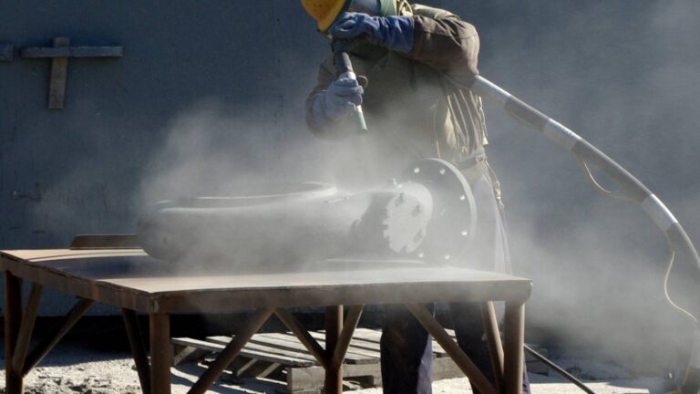 person sand blasting a metal pipe on a wooden table from a distance